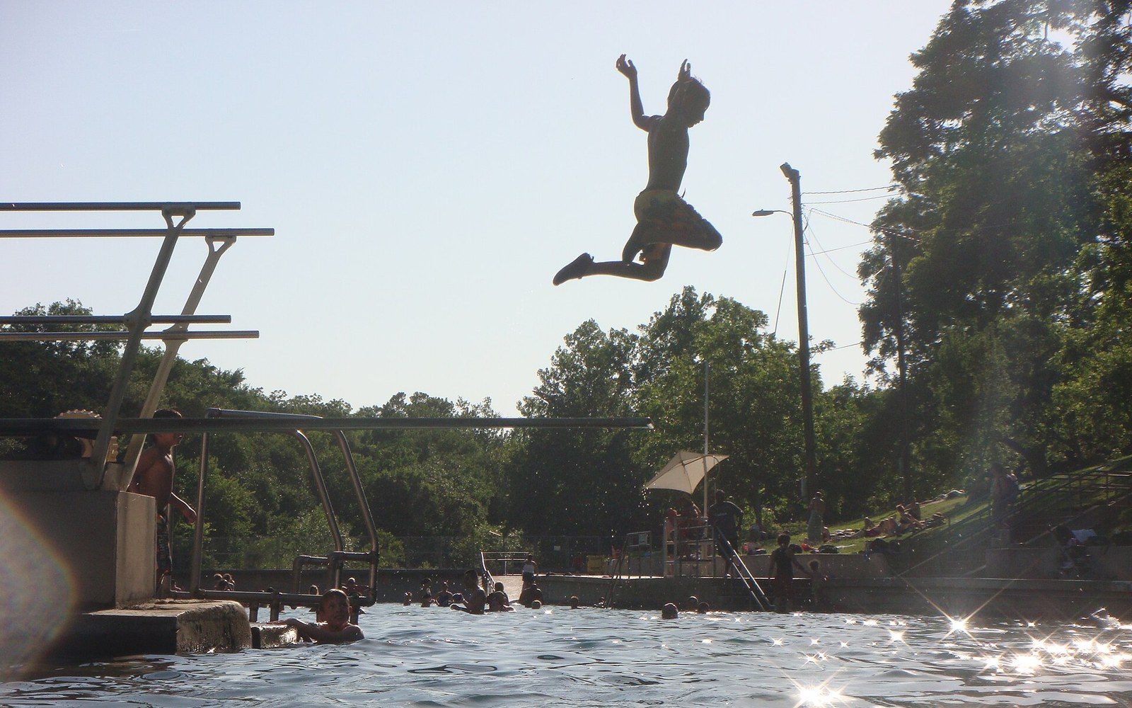 Barton Springs Pool viewed from the diving board toward the dam, with a swimmer and Zilker Park trees behind