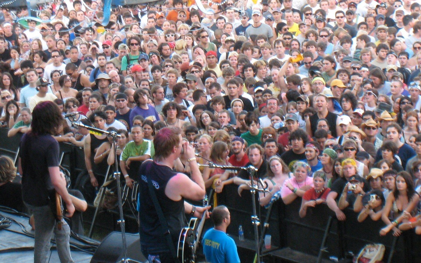 Musicians on stage play guitars facing a dense crowd of festival attendees