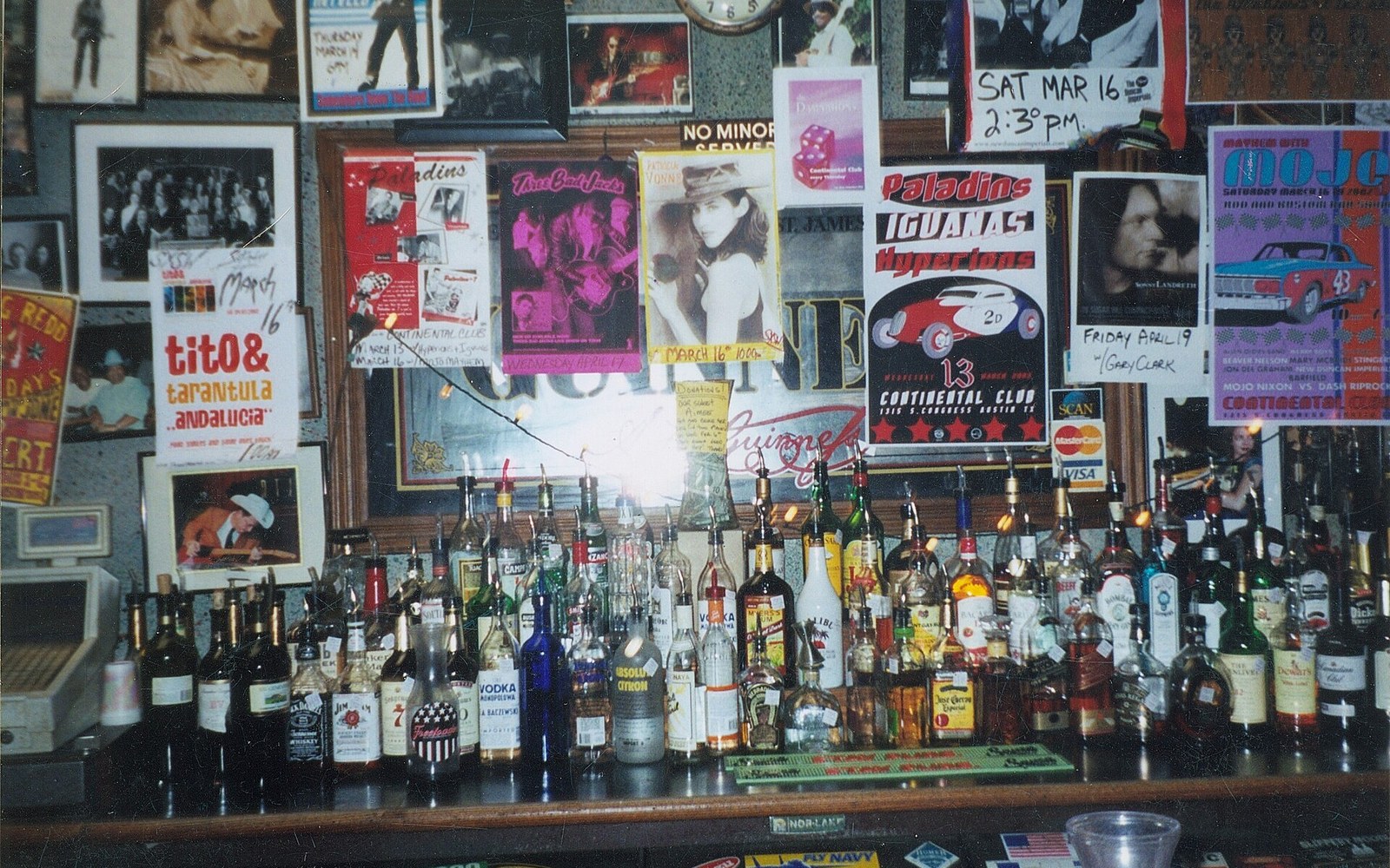 Bar shelf lined with liquor bottles beneath a wall covered in old concert posters