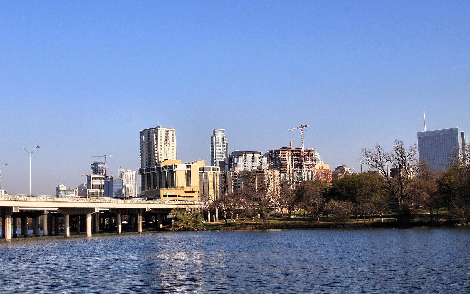 Downtown skyline with construction cranes rises beyond a bridge and riverside park