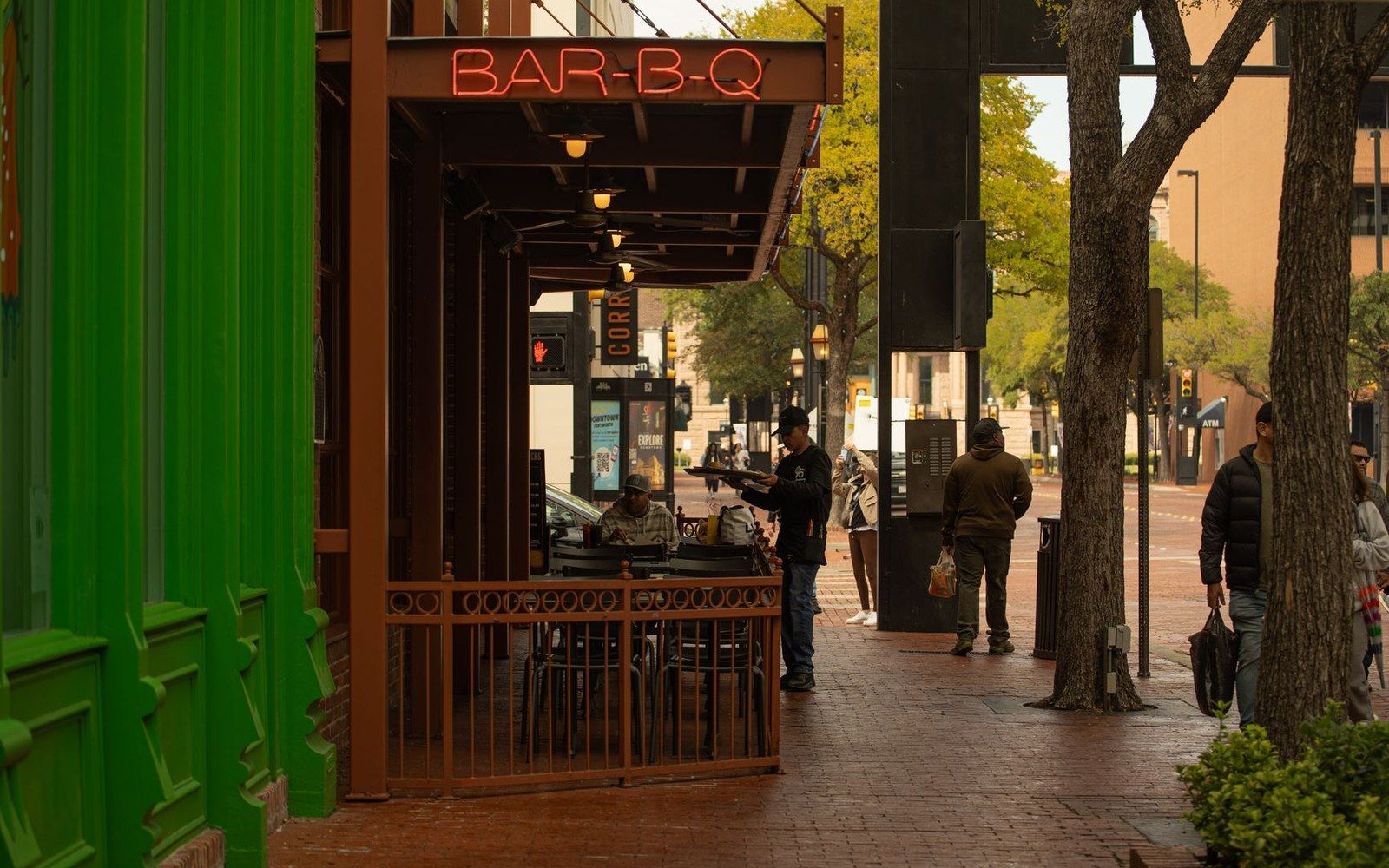 A neon "Bar-B-Q" sign glows above a sidewalk patio on a brick-paved city street