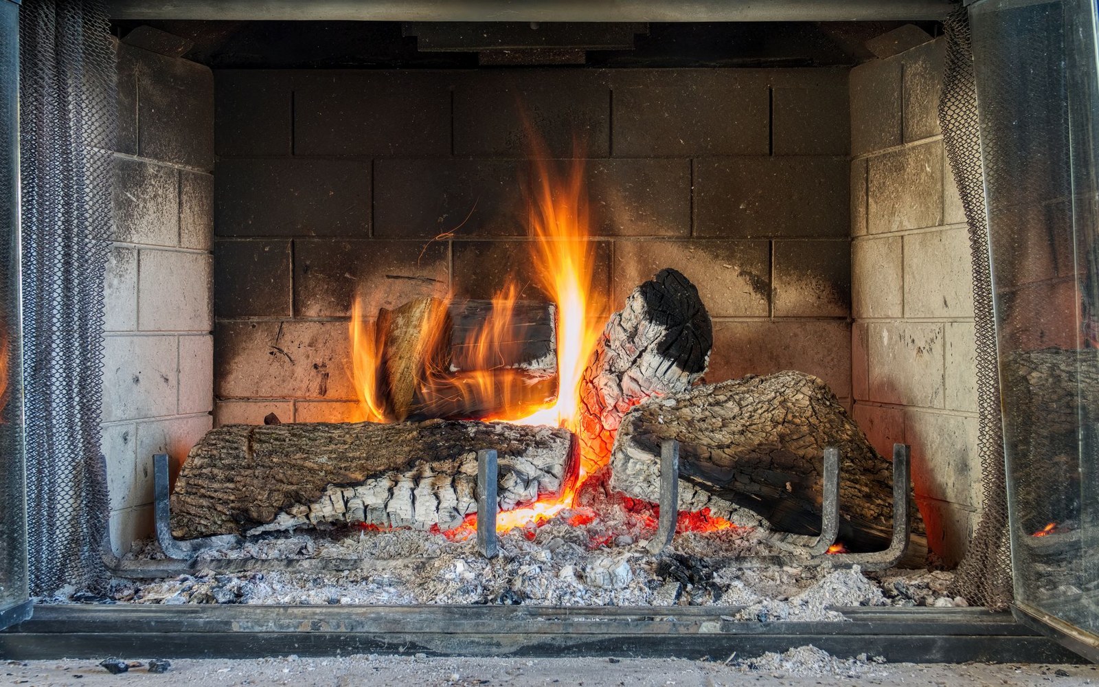 Burning logs and glowing embers inside a brick fireplace with mesh curtain screens