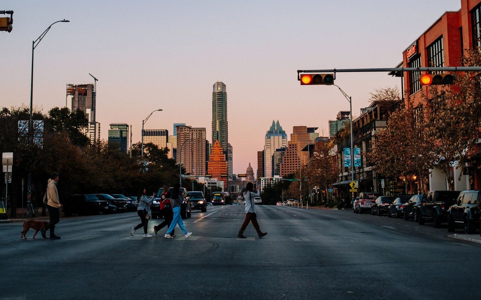 Pedestrians cross a downtown street at dusk with the Austin skyline and capitol dome behind them