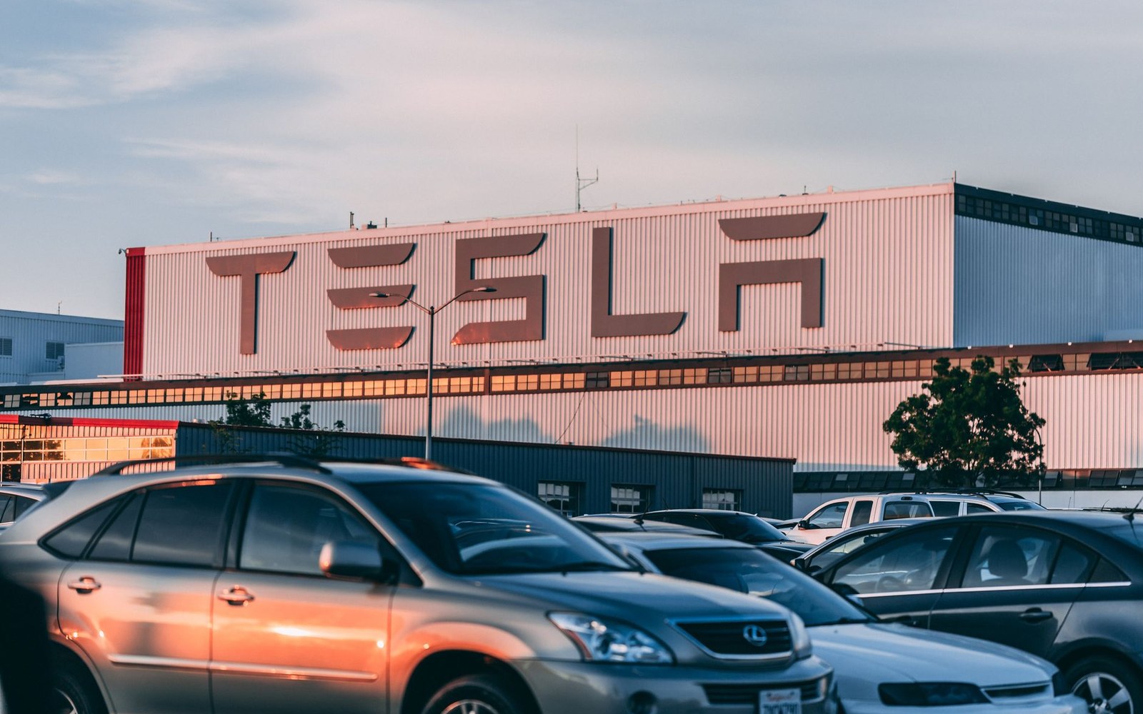 Parked cars in a lot outside a large Tesla factory building with rooftop logo