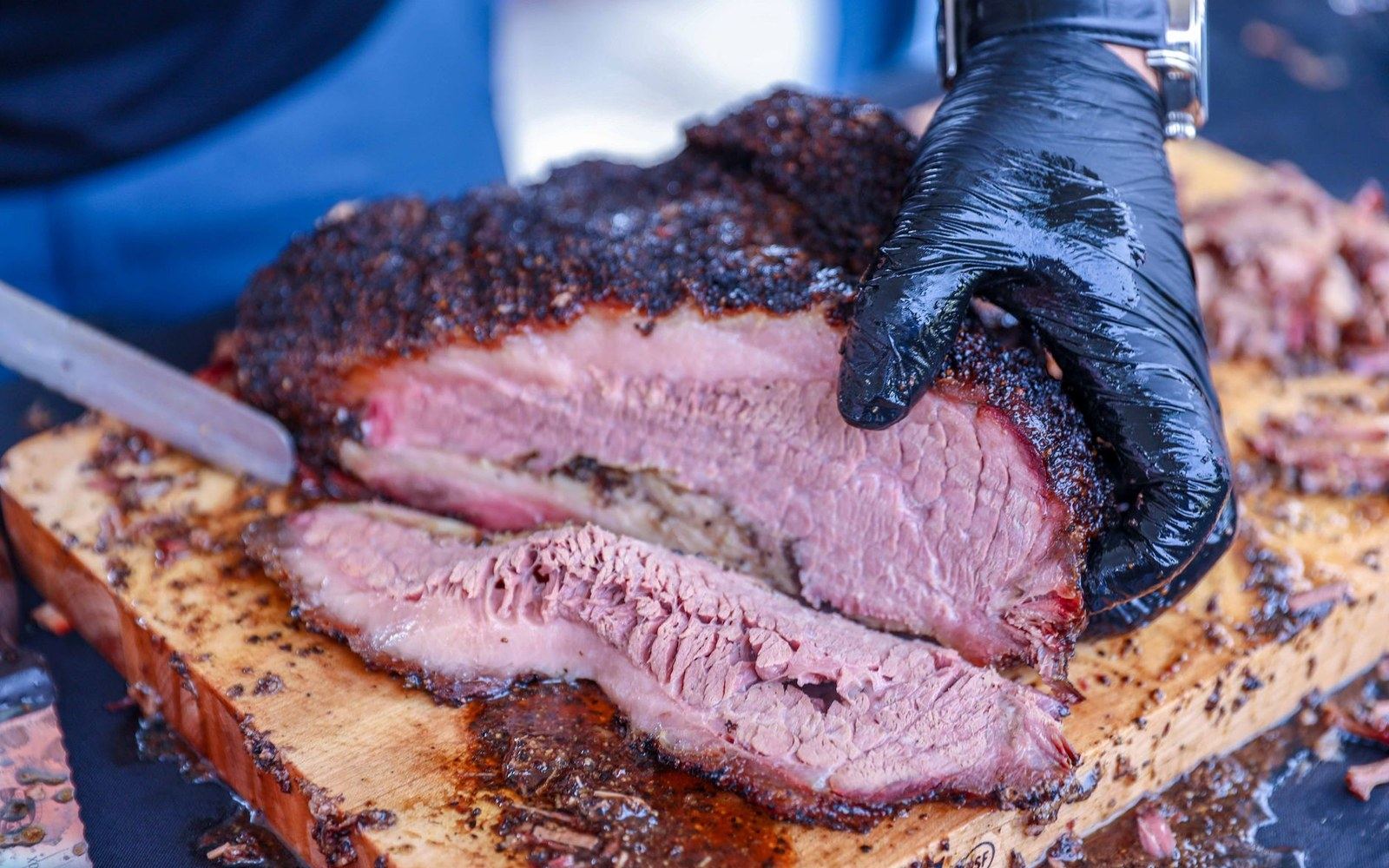 Adult pitmaster's gloved hands lifting a brisket with tongs from an open brick pit, post oak smoke visible, no face shown